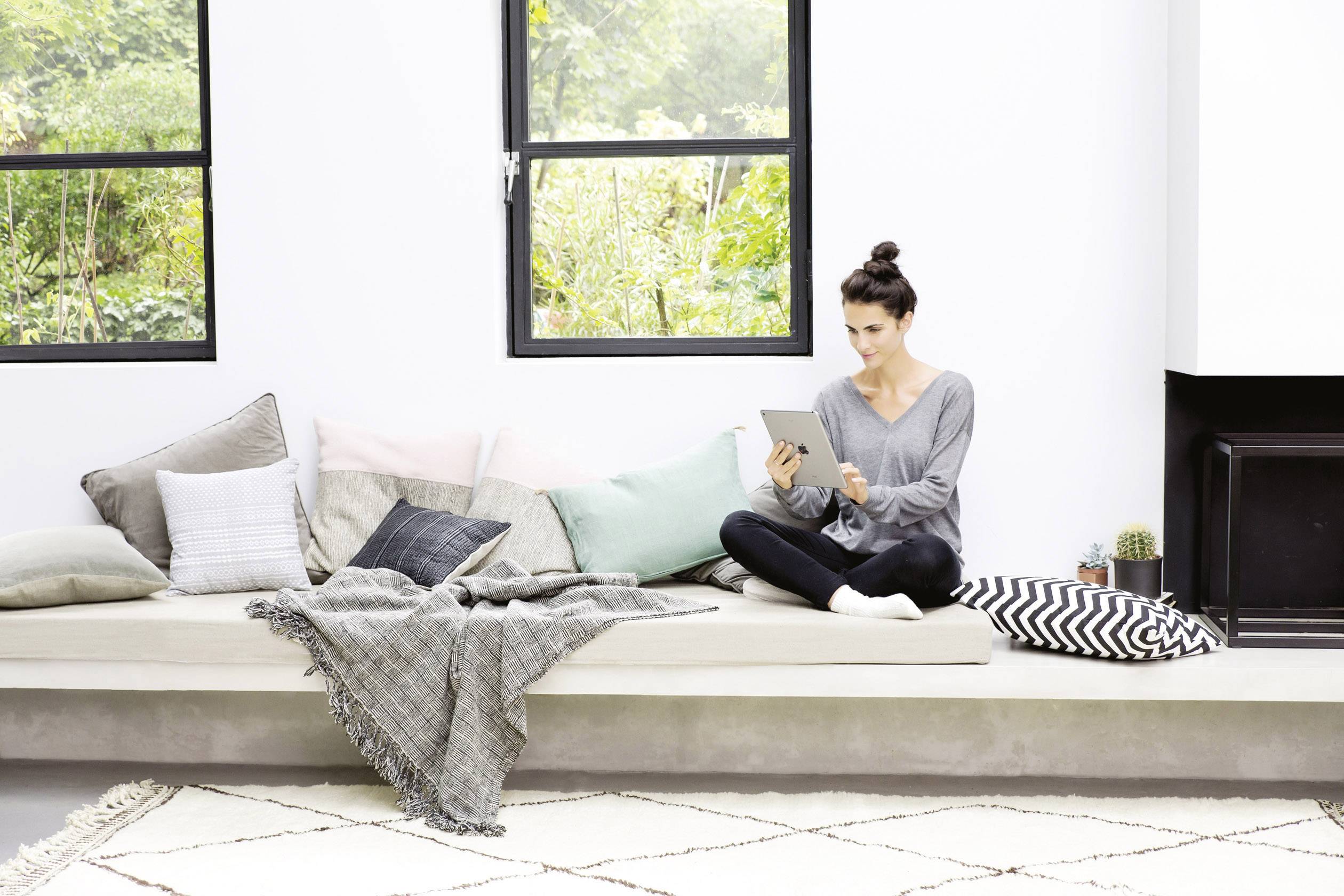 Een vrouw zit gezellig op een grote moderne bank met veel kussens en leest op haar tablet. Op de achtergrond zijn ramen.