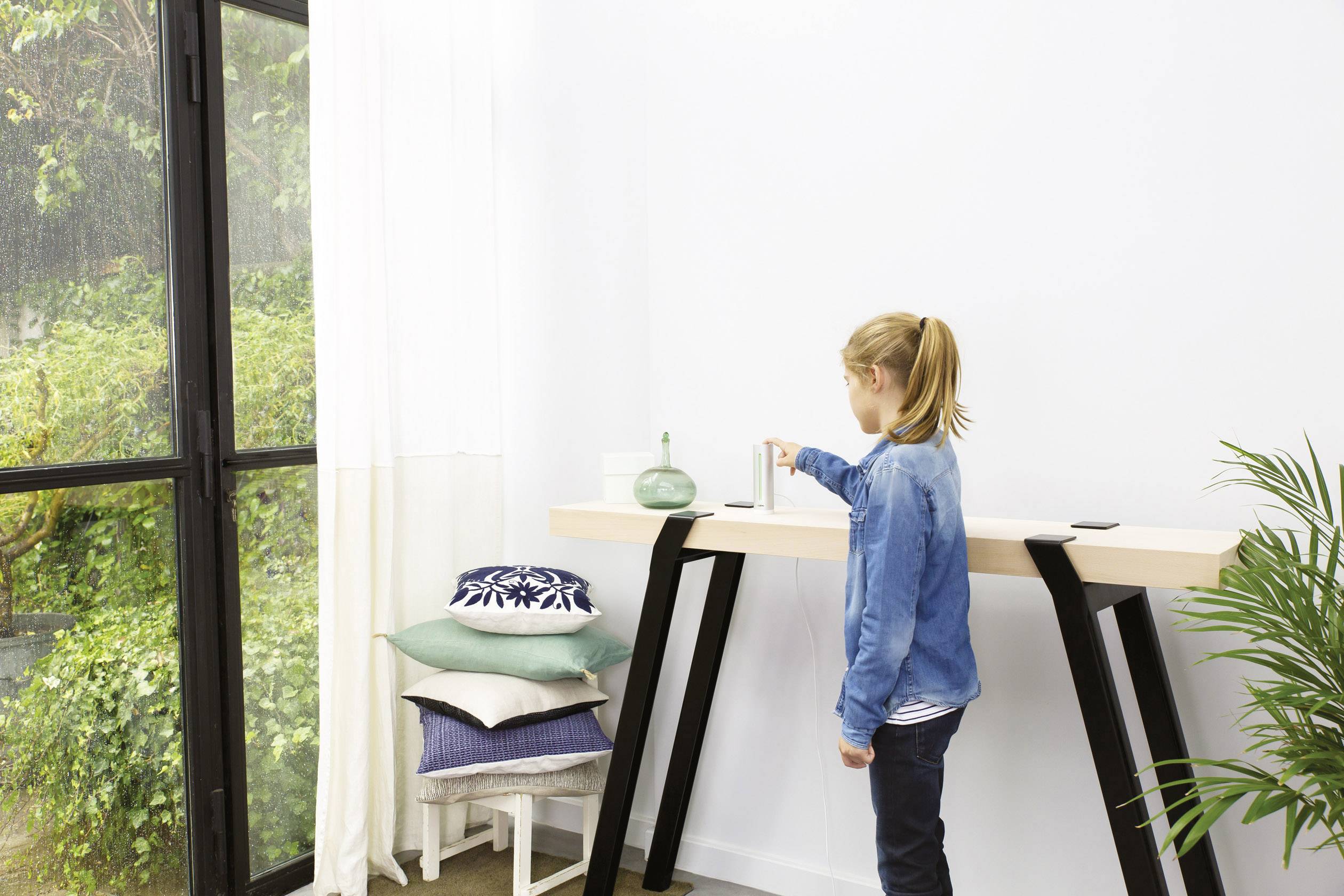 Een kind staat naast een hoge tafel en zet een glazen container erop. De ruimte is licht door een groot raam met zicht op planten.