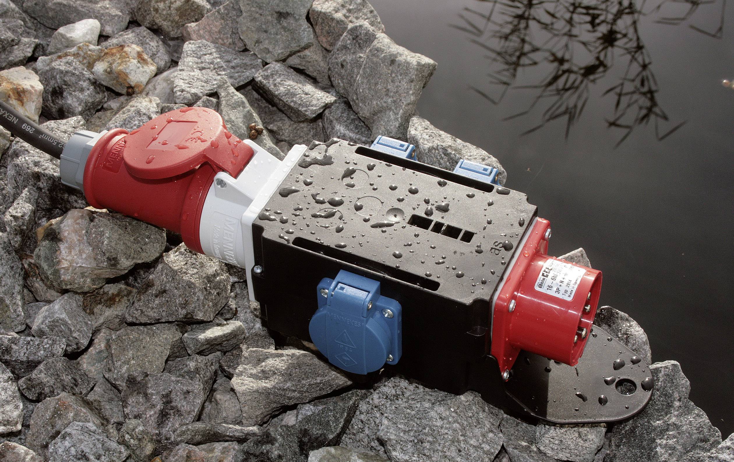 Een zwarte, rechthoekige elektrische verdeelkast met rode en blauwe stopcontacten ligt op natte stenen aan de oever van een waterpartij.