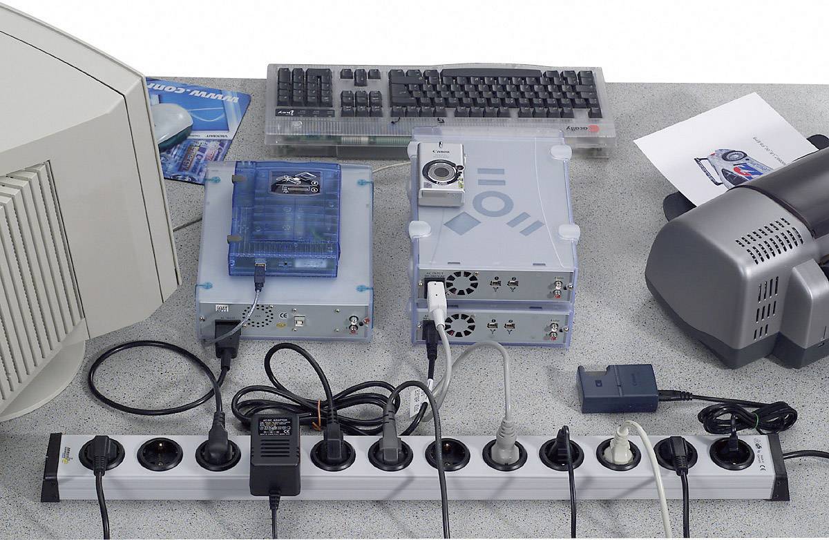 Een bureau met een monitor, toetsenbord, elektronische apparaten en meerdere kabels die zijn aangesloten op een stekkerdoos.