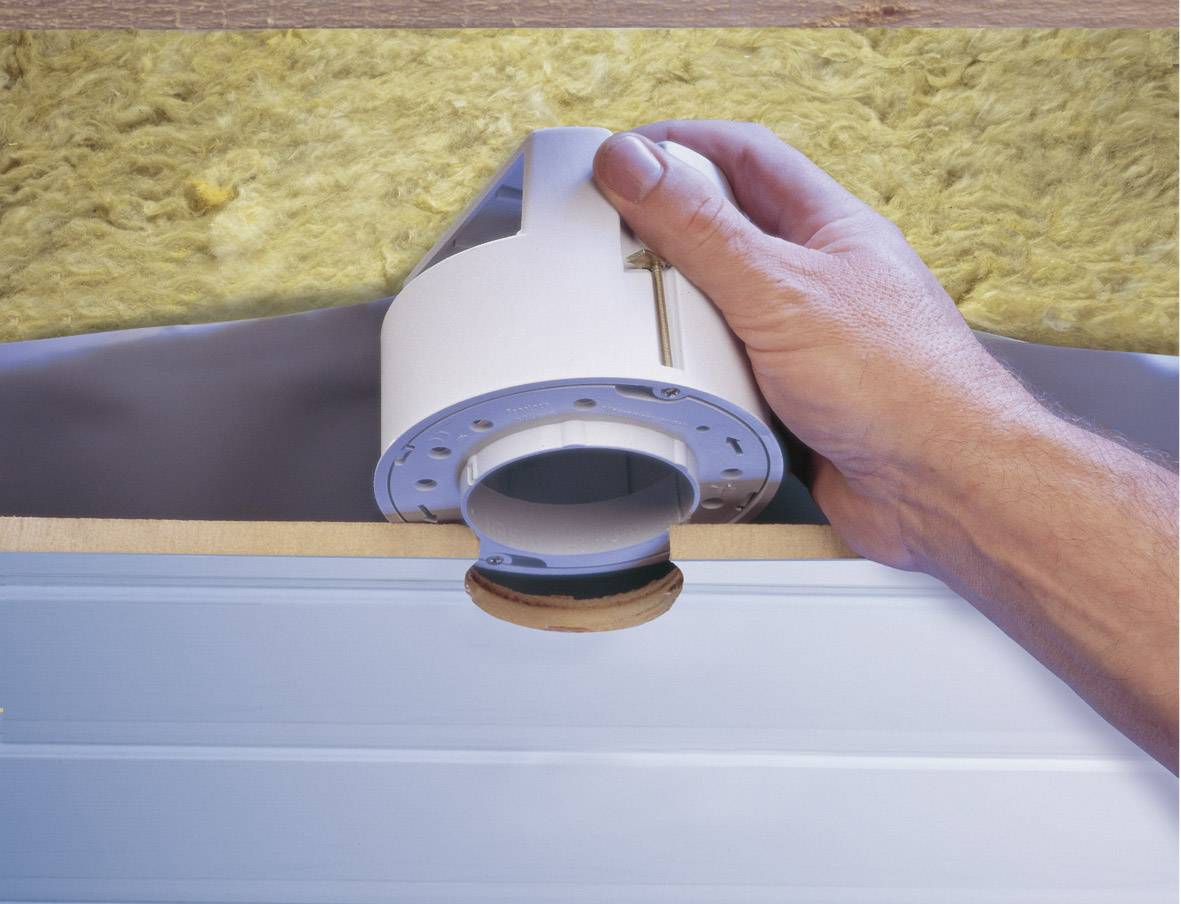 Een hand houdt een rond inbouwelement aan het plafond, nabij isolatiemateriaal. Installatie van een verlichtingsarmatuur.