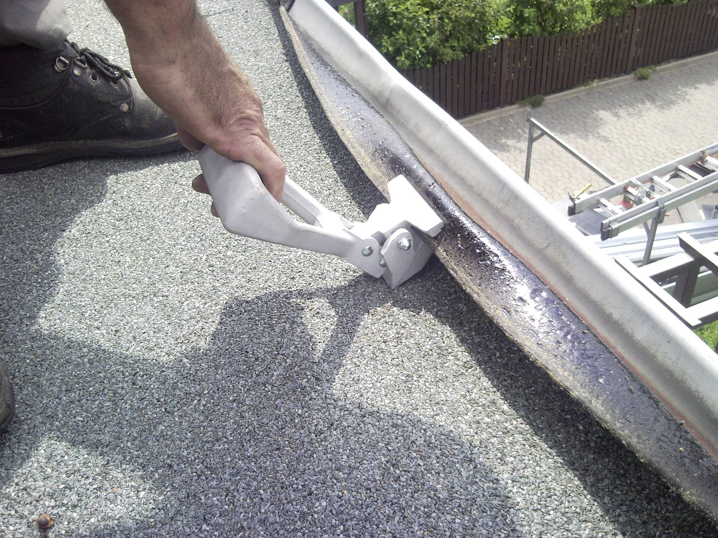 Een persoon bevestigt een metalen onderdeel aan een dakrand op een met grind bedekt plat dak. Op de achtergrond is een ladder zichtbaar.