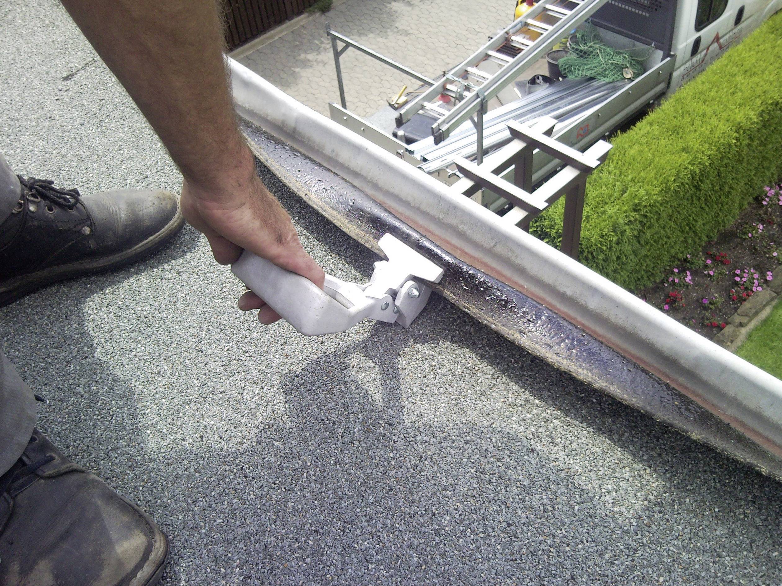 Een persoon reinigt een dakgoot met een gereedschap. Op de achtergrond zijn een ladder en een bestelwagen zichtbaar.