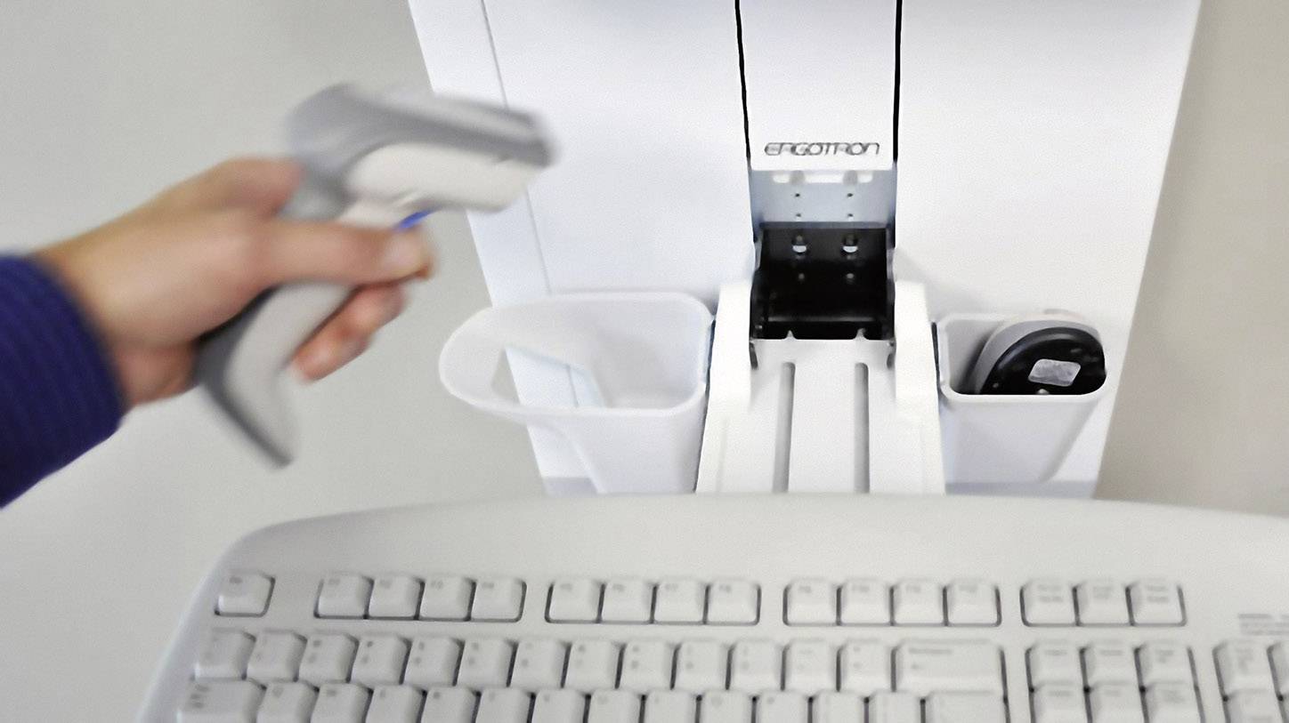 Een hand houdt een barcodescanner vast boven een wit toetsenbord op een bureau. Rechts bevinden zich opbergvakken voor kantoorbenodigdheden.