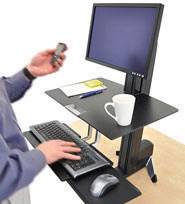 Persoon werkt aan een in hoogte verstelbare sta-bureau met monitor, toetsenbord en muis. Persoon houdt een smartphone in de hand.