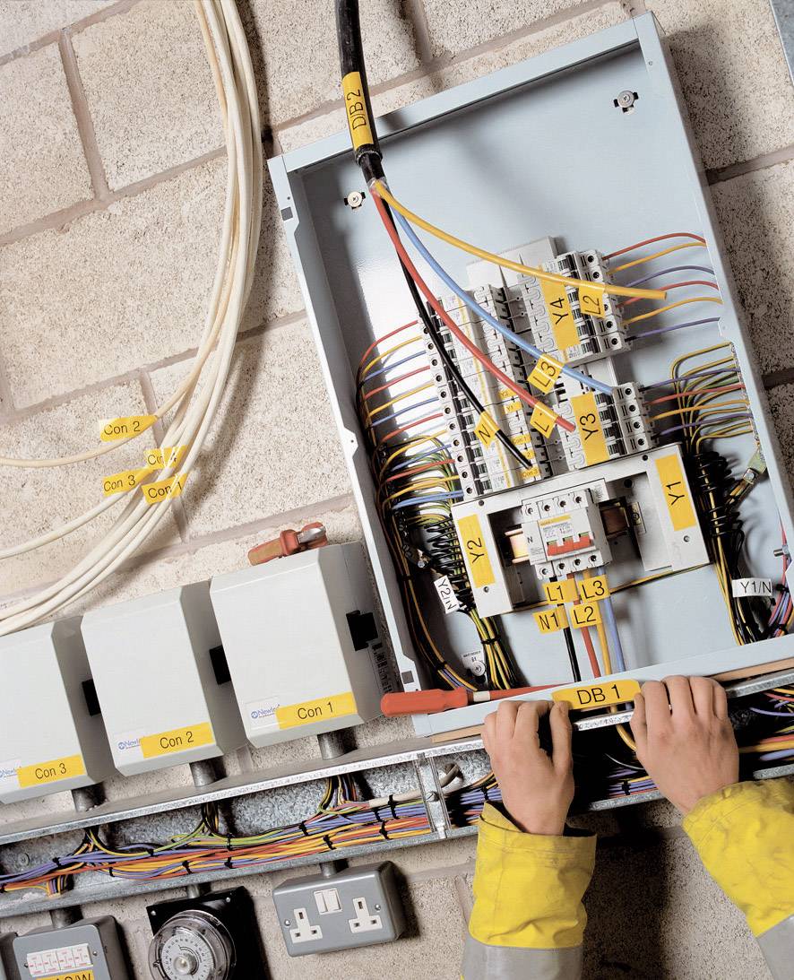 Een persoon werkt aan een geopende elektrische schakelkast. Verschillende draden zijn voorzien van labels om de verbindingen aan te duiden.