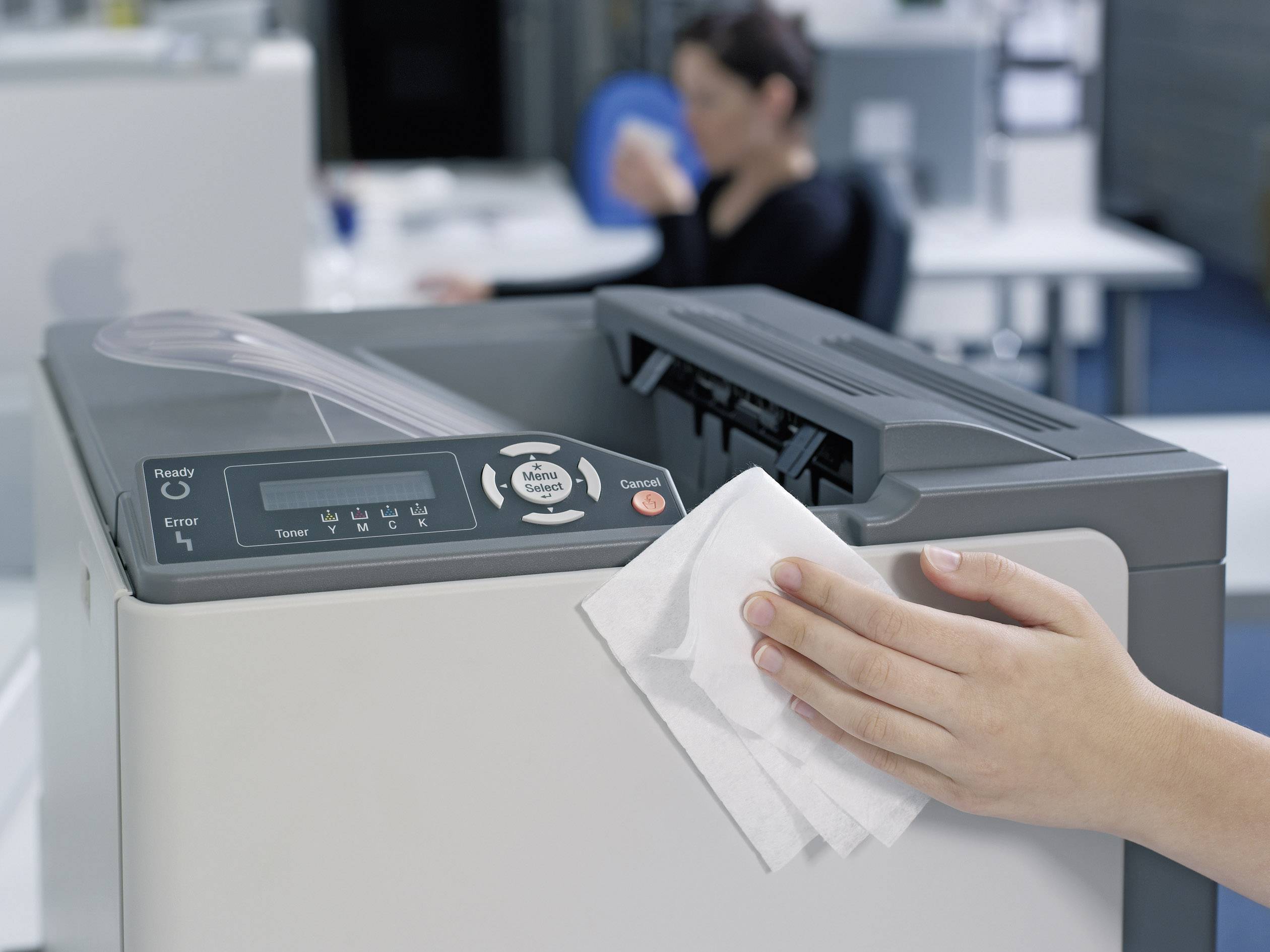 Een persoon reinigt een printer in het kantoor met een doek, terwijl op de achtergrond wazige kantoormeubels en een werkende persoon te zien zijn.