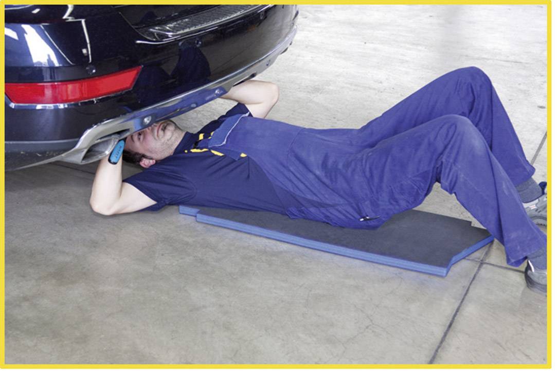 Persoon in blauwe werkkleding ligt op een mat onder de achterkant van een zwarte auto en inspecteert deze in een werkplaats.