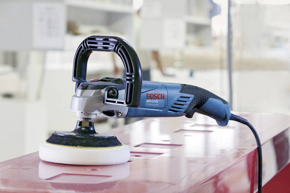 Elektrische slijpmachine in een werkplaatsomgeving op een metalen tafel. Het apparaat is blauw met een zwarte handgreep en een wit slijpschijf.