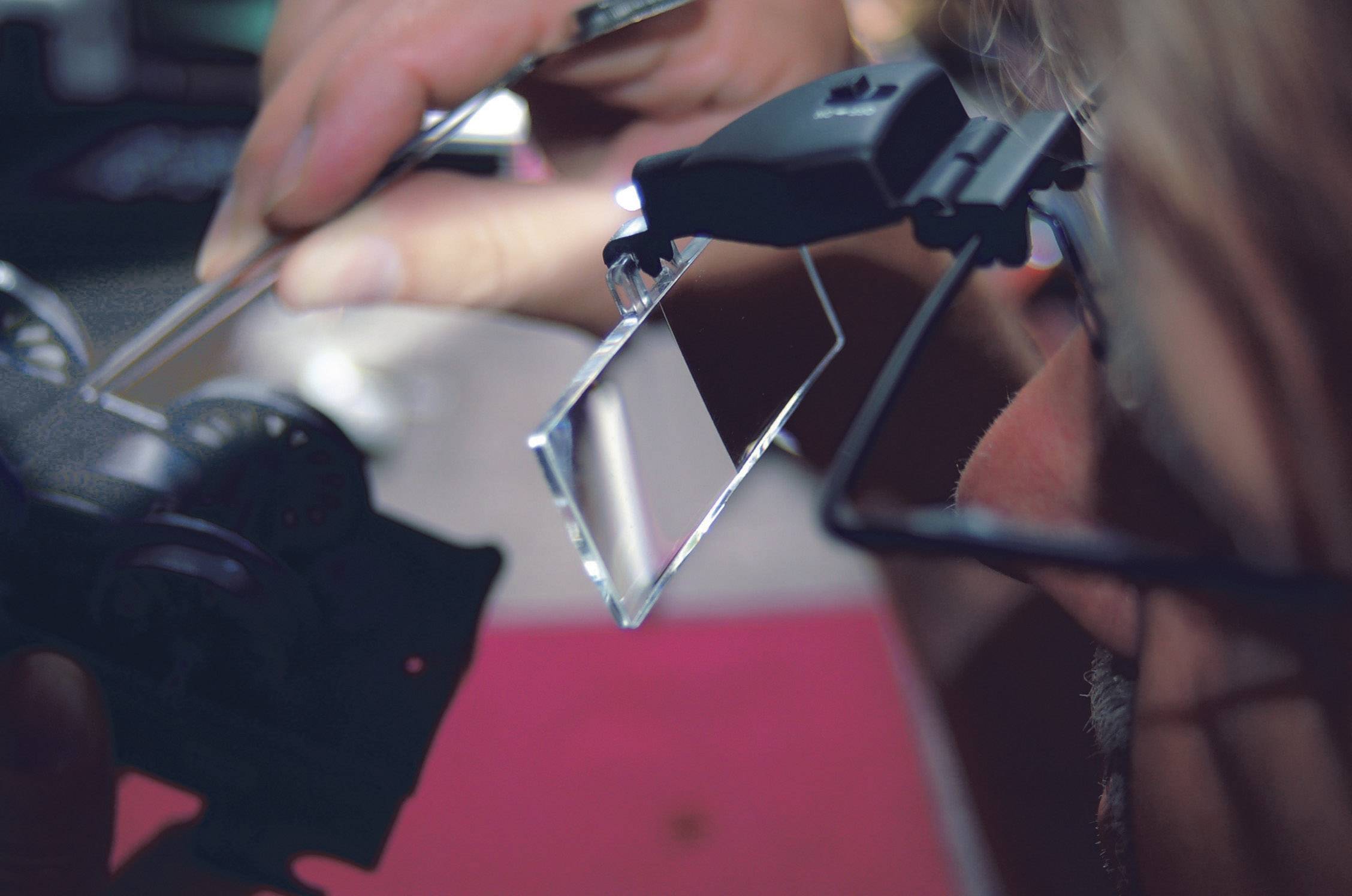 Een persoon inspecteert een klein technisch apparaat met een vergrootglas. Handen en gereedschap zijn zichtbaar terwijl gedetailleerde onderdelen nauwkeurig worden onderzocht.