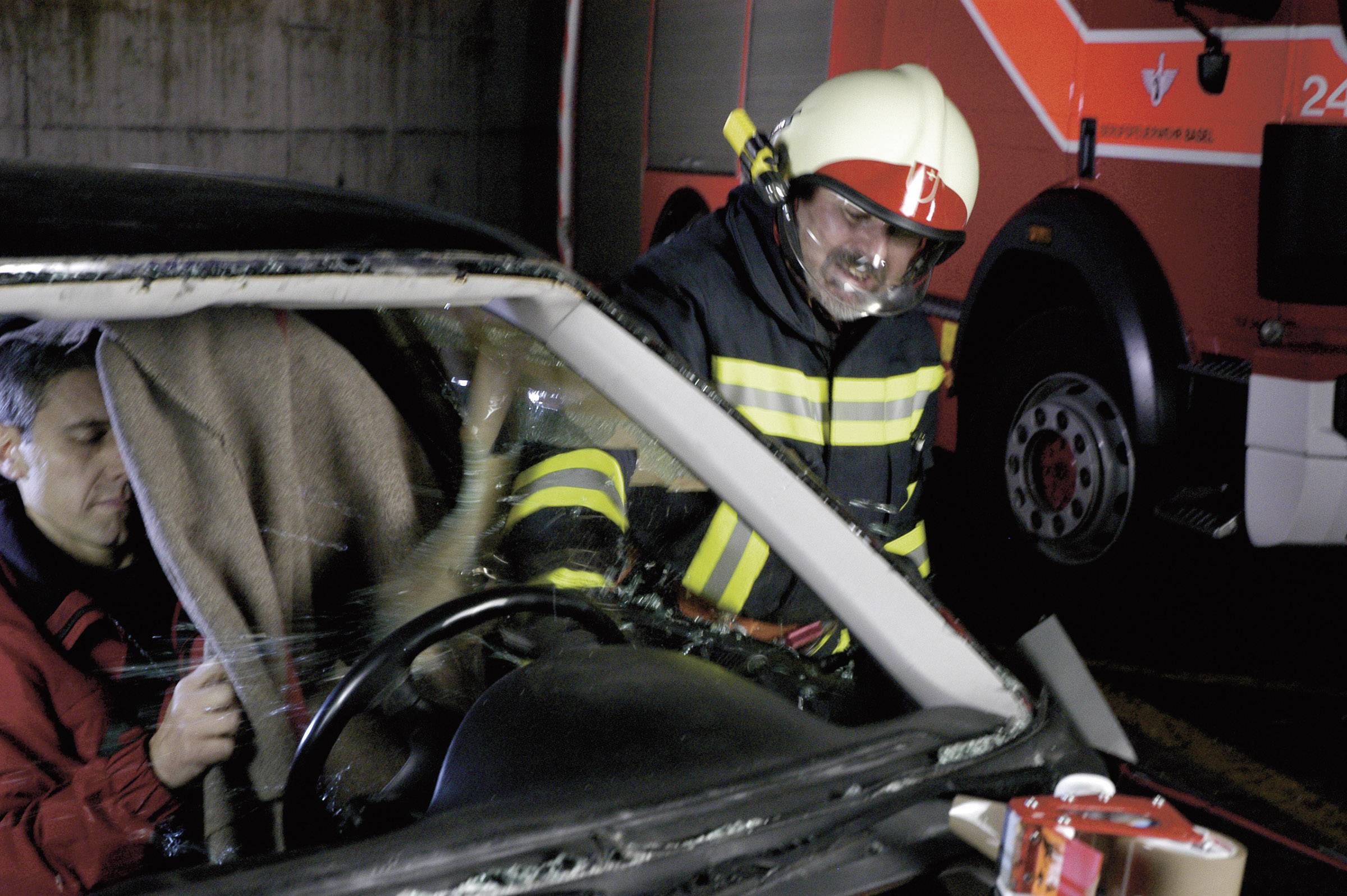 Een brandweerman redt een persoon uit een auto na een ongeluk. Een brandweerwagen is op de achtergrond zichtbaar.