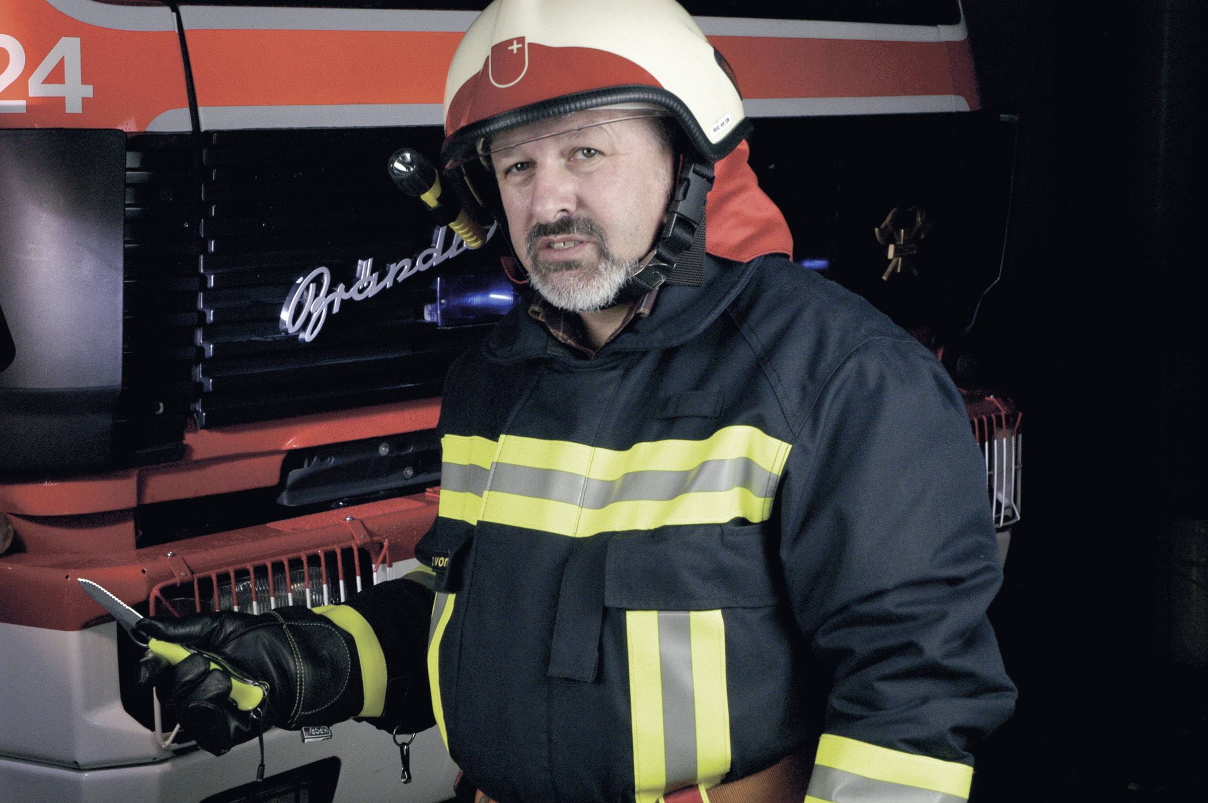 Een brandweerman in volledige uitrusting staat voor een brandweerwagen en houdt een portofoon vast.