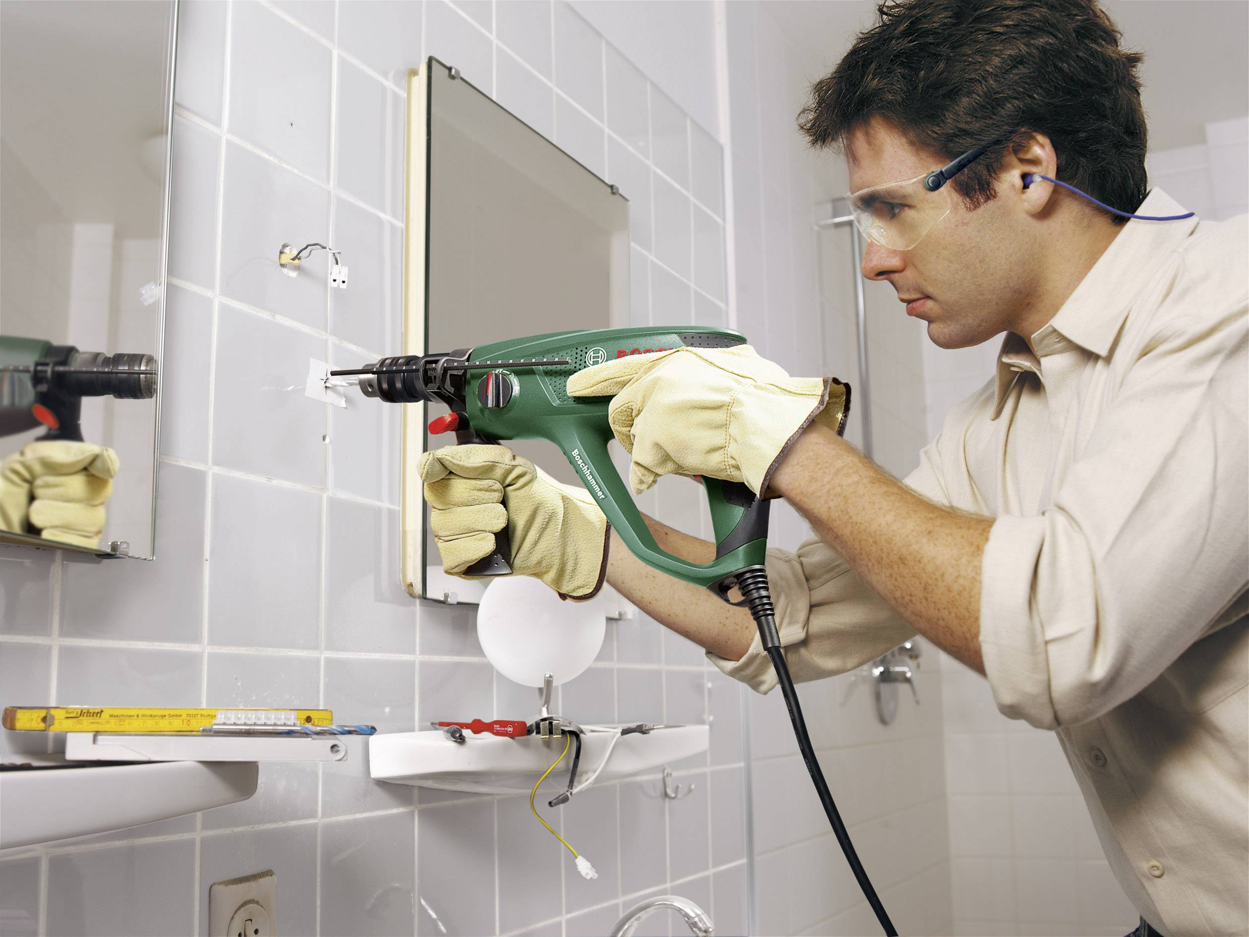 Een man met veiligheidsbril en handschoenen boort een gat in een badkamertegel boven een wastafel. Gereedschappen liggen klaar.