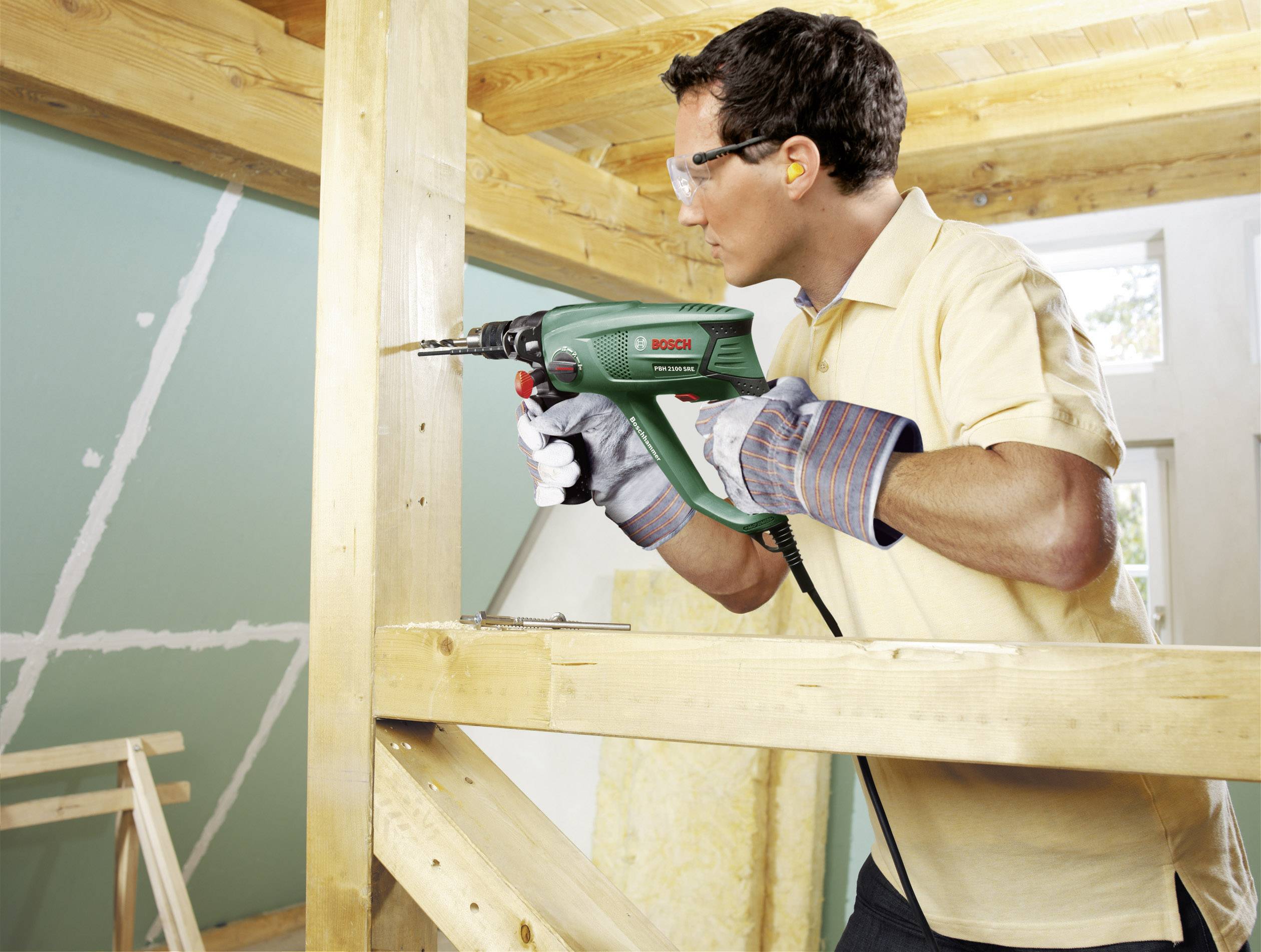 Man met veiligheidsbril en handschoenen boort een gat in een houten paal in een ruimte die wordt gerenoveerd.