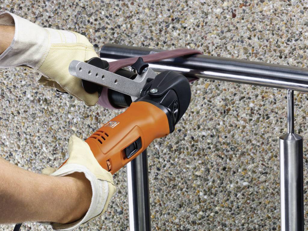 Een persoon slijpt een metalen leuning met een haakse slijper, waarbij hij handschoenen draagt voor veiligheid. Op de achtergrond is een stenen muur zichtbaar.