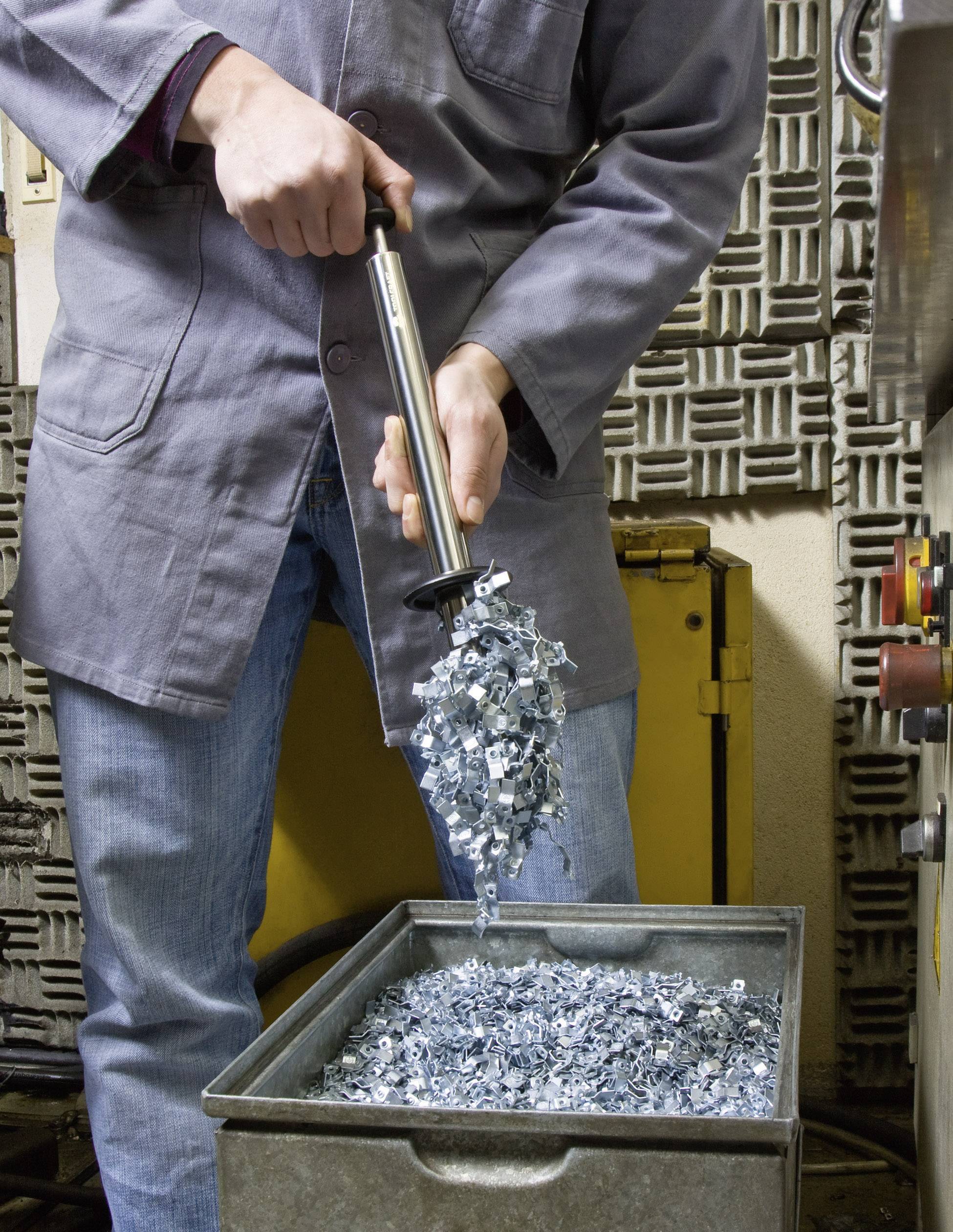 Een technicus in beschermende kleding houdt metalen onderdelen vast met een magneetstaaf boven een container. De onderdelen vallen van de magneet in de doos.