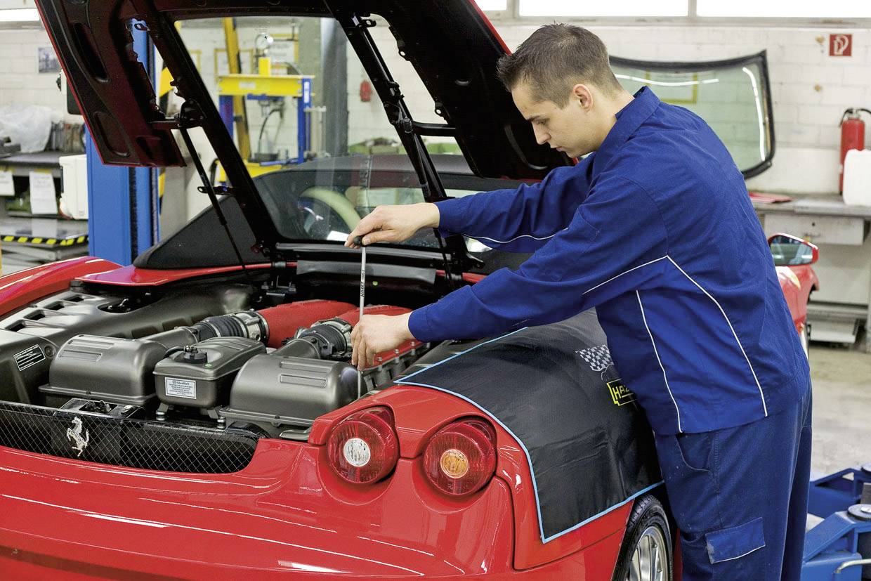Monteur in blauwe overall werkt aan de motor van een rode sportwagen in een werkplaats. De kofferbak staat open.