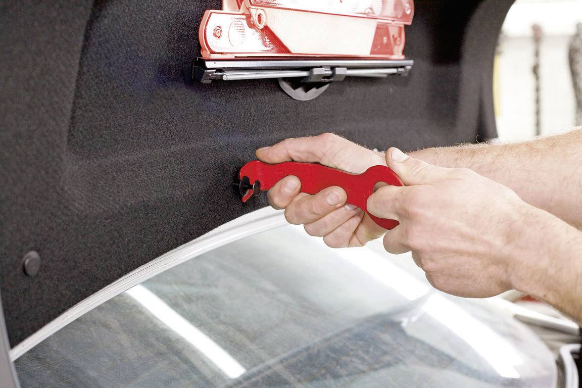 Een persoon gebruikt een rood gereedschap om een onderdeel in de kofferbak van een auto te bewerken. Een EHBO-doos is op de achtergrond zichtbaar.
