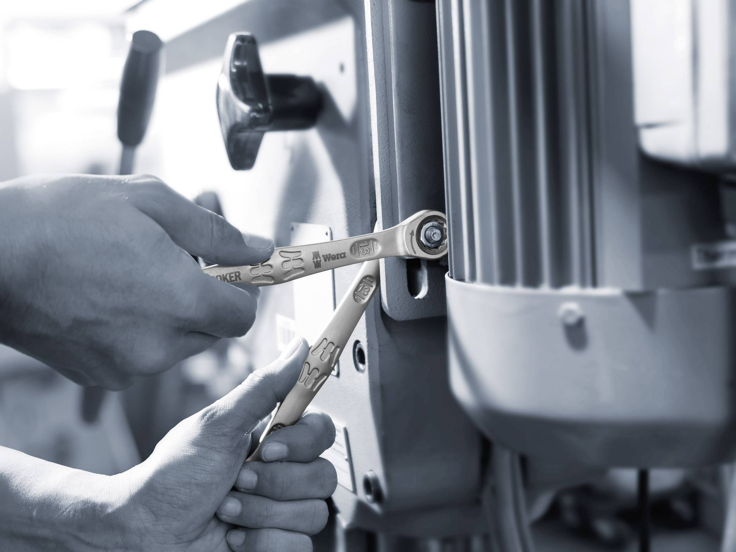 Twee handen gebruiken moersleutels om een machine te repareren. De focus ligt op het nauwkeurig vastdraaien van schroeven.