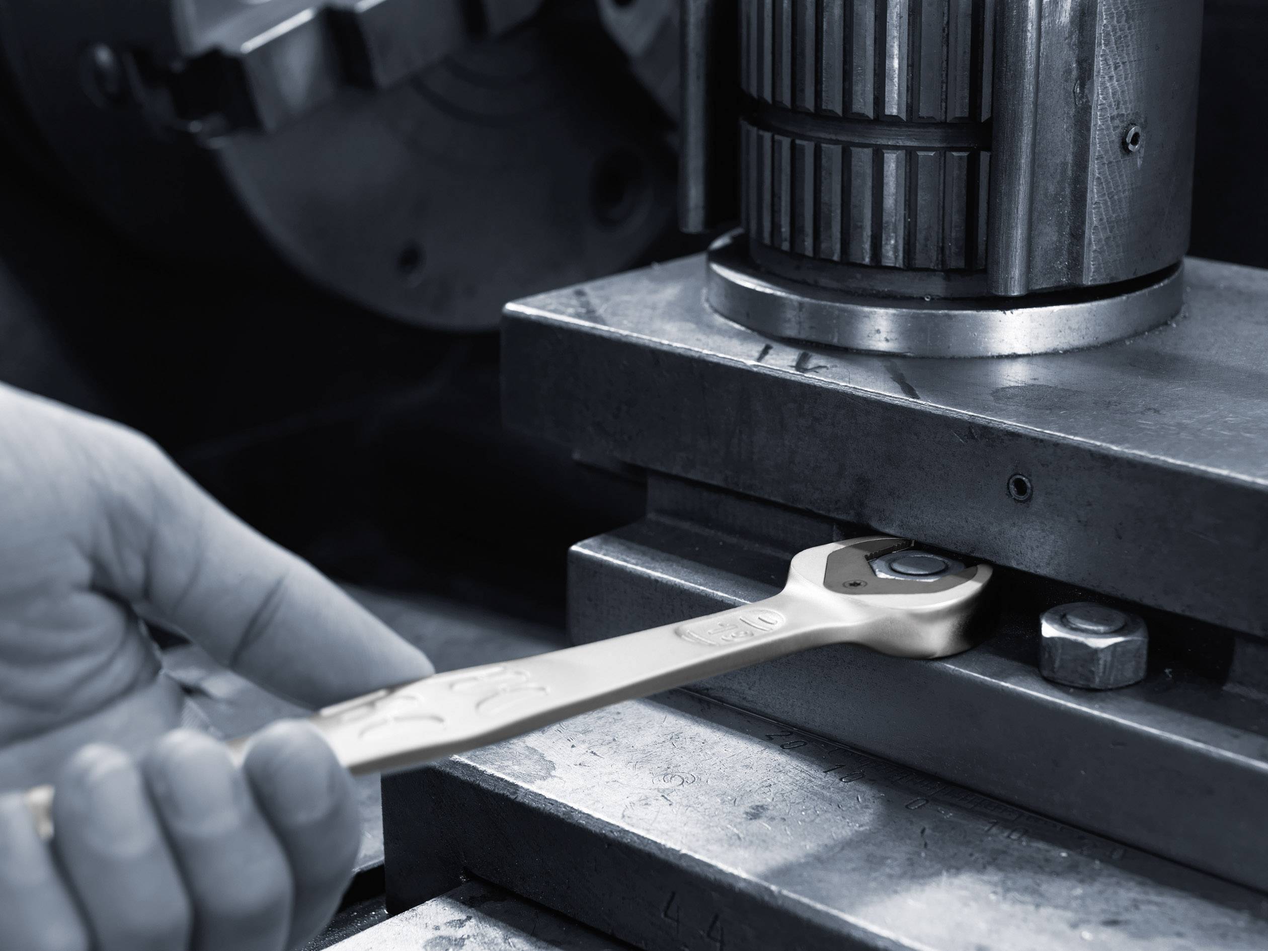 Een hand houdt een moersleutel vast aan een machine. De focus ligt op nauwkeurige afstelling en onderhoud van de machinedelen.