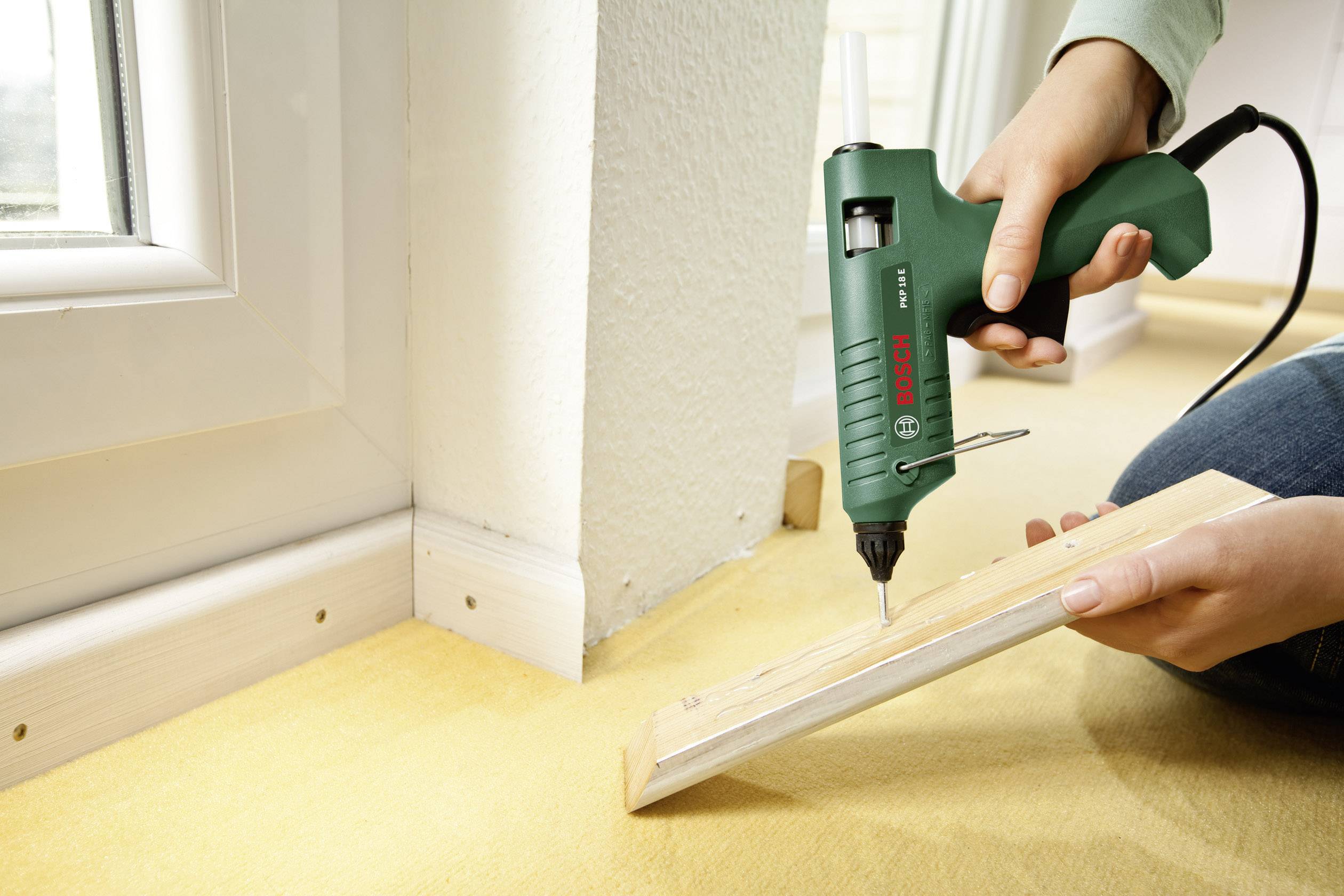 Een persoon gebruikt een groene lijmpistool om een stuk hout vast te maken in een hoek van een kamer. Huisdeur op de achtergrond.