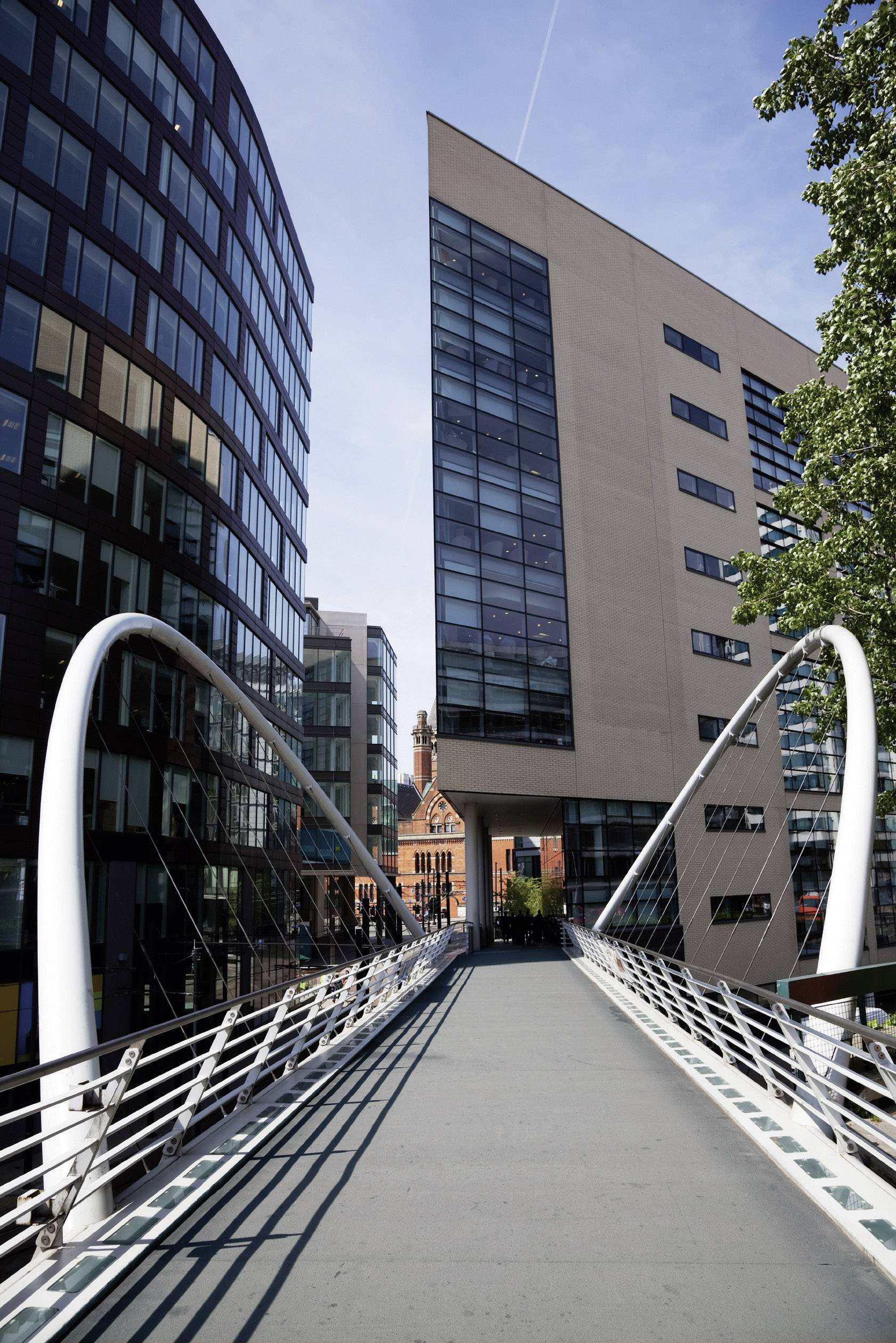 Een moderne voetgangersbrug loopt tussen hoge kantoorgebouwen door. Op de achtergrond is een rood bakstenen gebouw zichtbaar.