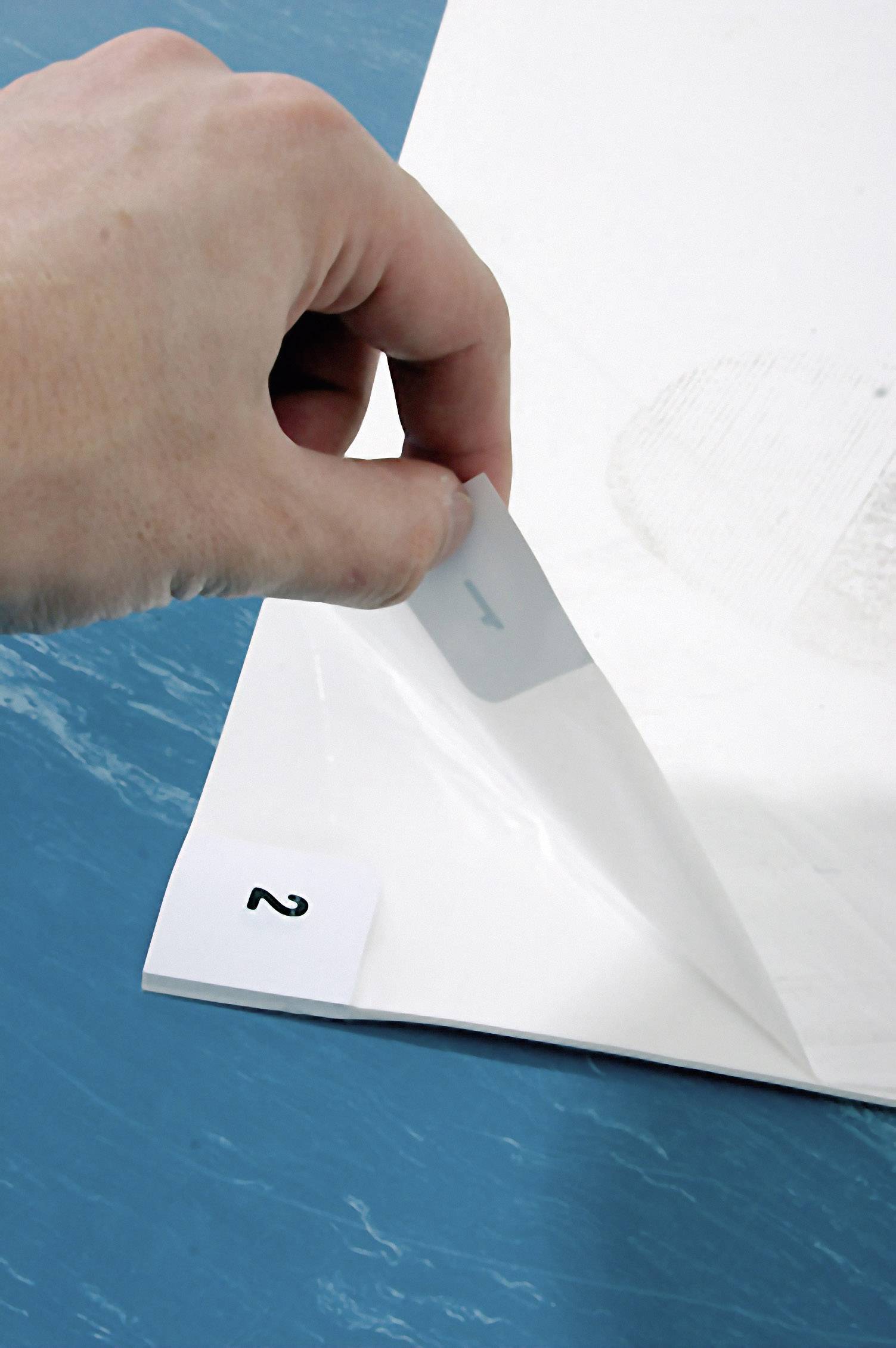 Hand heeft een transparante folie van een vel papier met het getal '2' aan de rand, op een blauwe achtergrond.