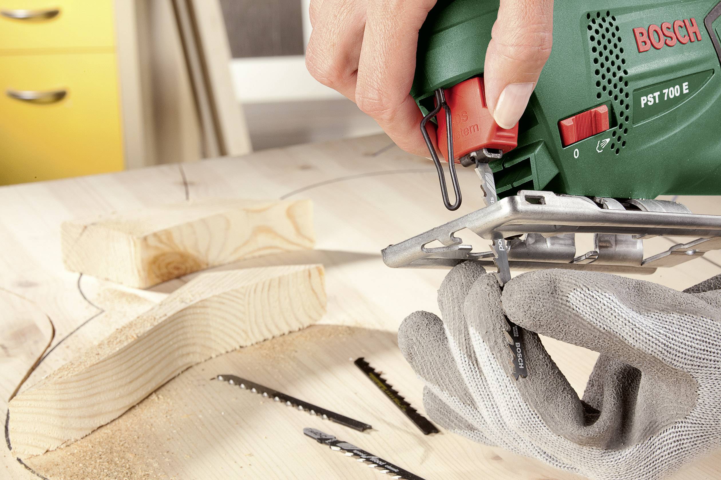 Een persoon zaagt een gebogen vorm uit een houten plank met een groene decoupeerzaag, terwijl hij grijze veiligheidshandschoenen draagt.