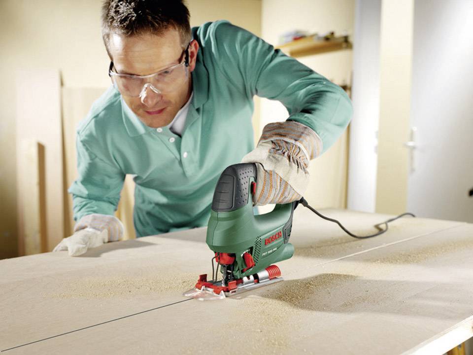 Man met veiligheidsbril en handschoenen bedient een groene decoupeerzaag die een houten plaat aan het doorsnijden is.