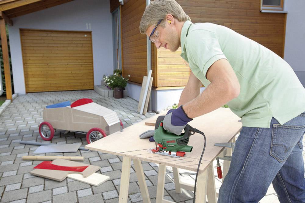 Een man met veiligheidsbril en handschoenen zaagt hout met een decoupeerzaag op een werkbank in een binnenplaats. Op de achtergrond staat een rood en beige speelgoedautootje voor kinderen.