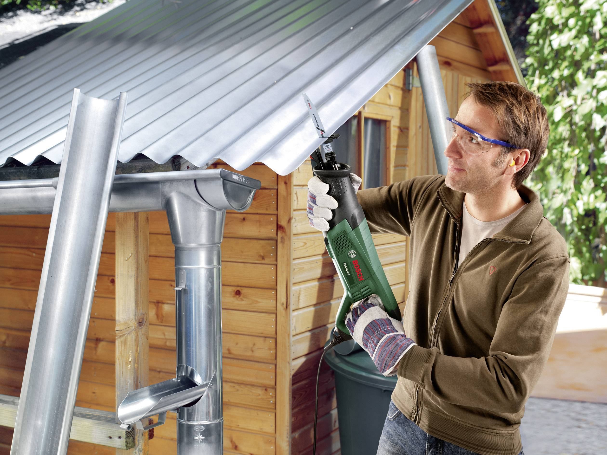 Een man werkt met een reciprozaag op een golfplaten dak naast een houten schuur. Hij draagt een veiligheidsbril en handschoenen.