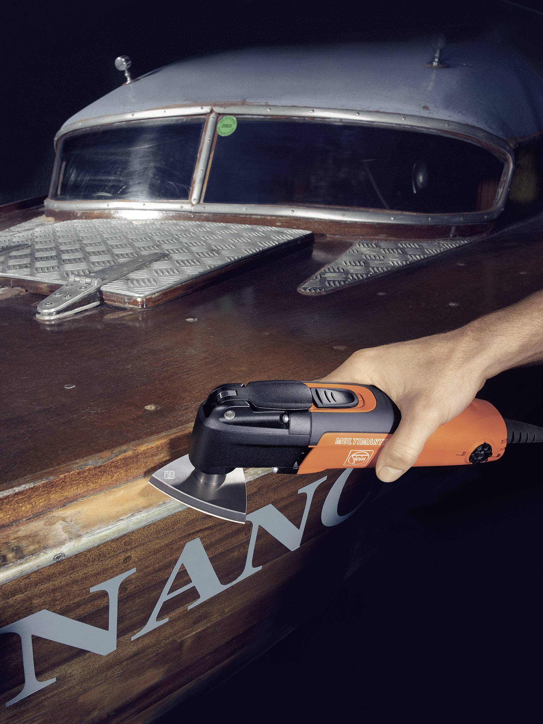 Een persoon schuurt met een elektrische schuurmachine de lak van een houten boot. Op de achtergrond is het dek van de boot zichtbaar.