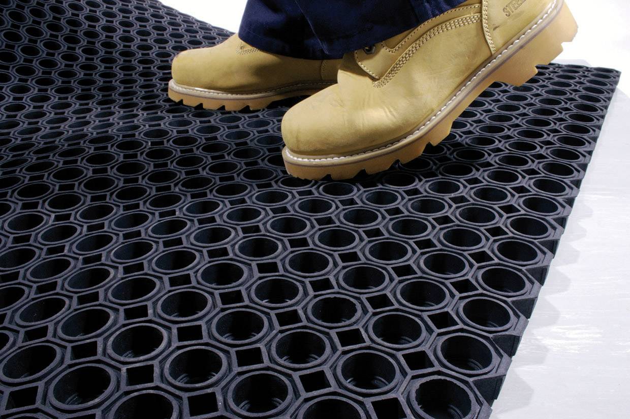 Een paar gele werkschoenen staat op een zwarte, slipvrije vloermat met honingraatvormige gaten die voor stevige grip zorgen.