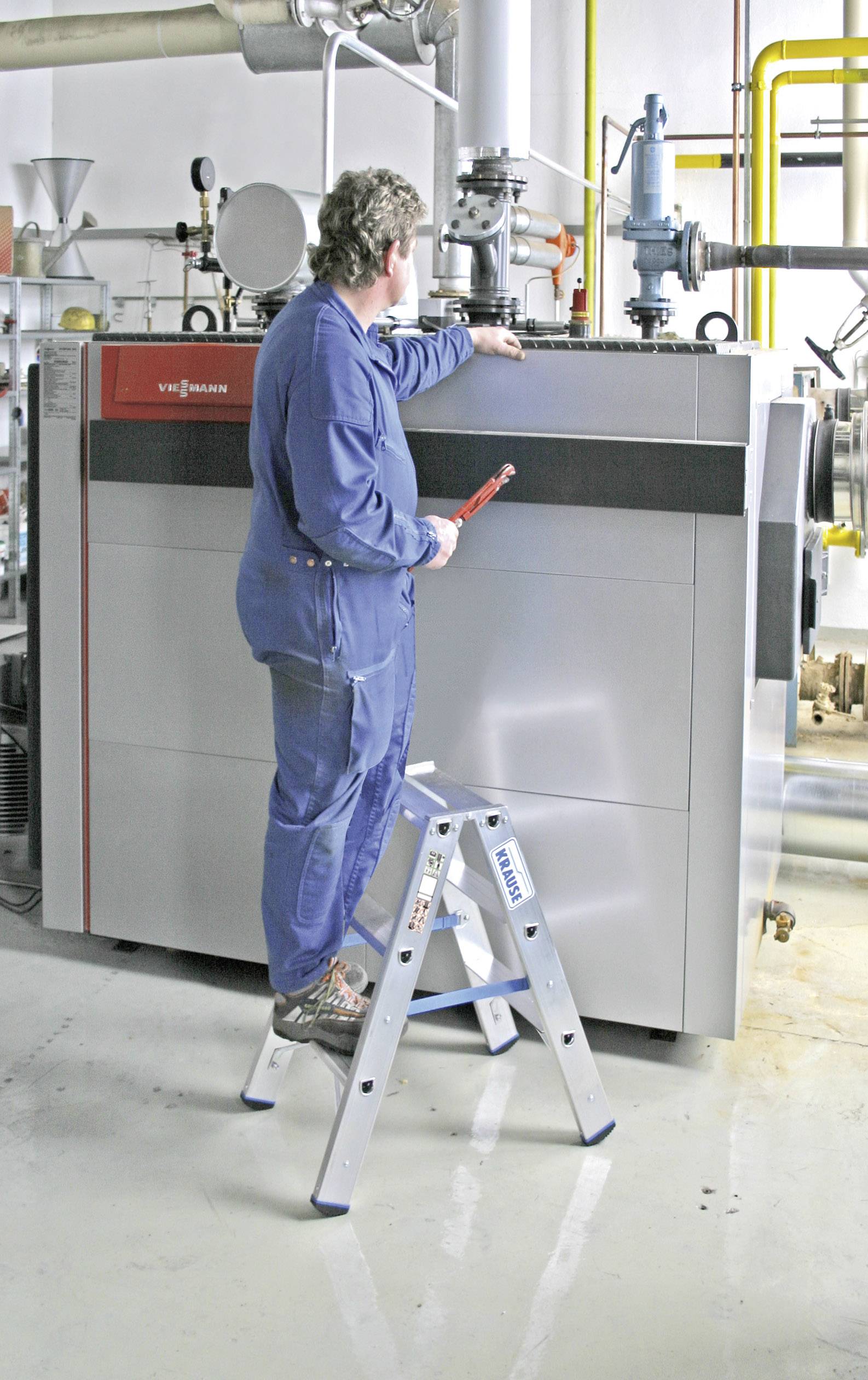 Een technicus in blauwe werkkleding staat op een ladder en werkt aan een grote industriële machine in een werkplaatsomgeving.