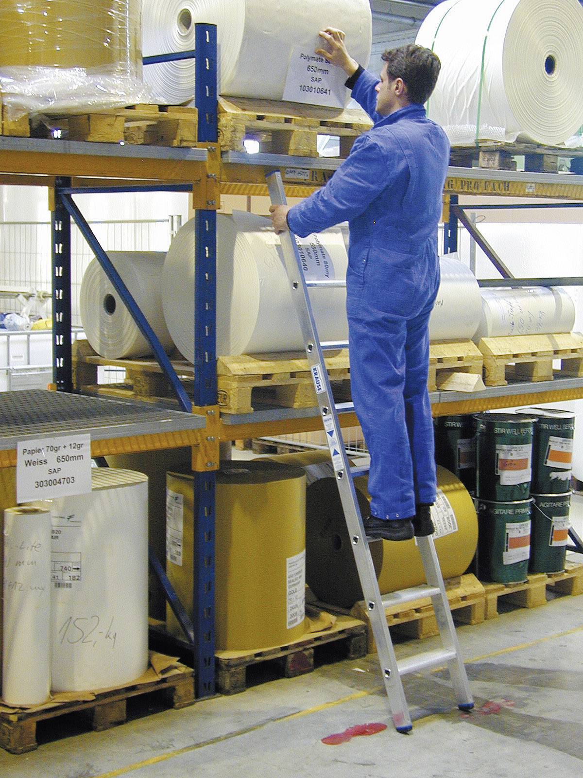 Een persoon in blauwe werkkleding staat op een ladder en controleert grote rollen papier of stof op een magazijnschap.