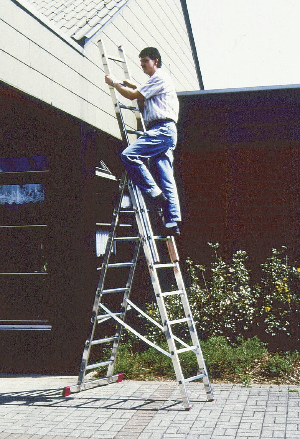 Een persoon klimt op een grote aluminium ladder die tegen een gebouw leunt. Hij/zij werkt in de buurt van het dak.