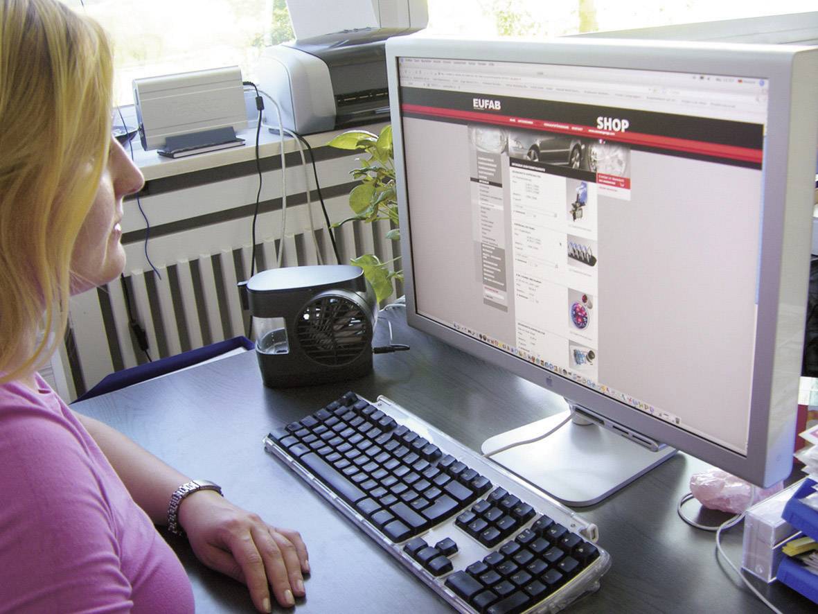 Een vrouw zit aan haar bureau en bekijkt een winkelwebsite op een computerscherm. Toetsenbord, muis en elektronica op de voorgrond.