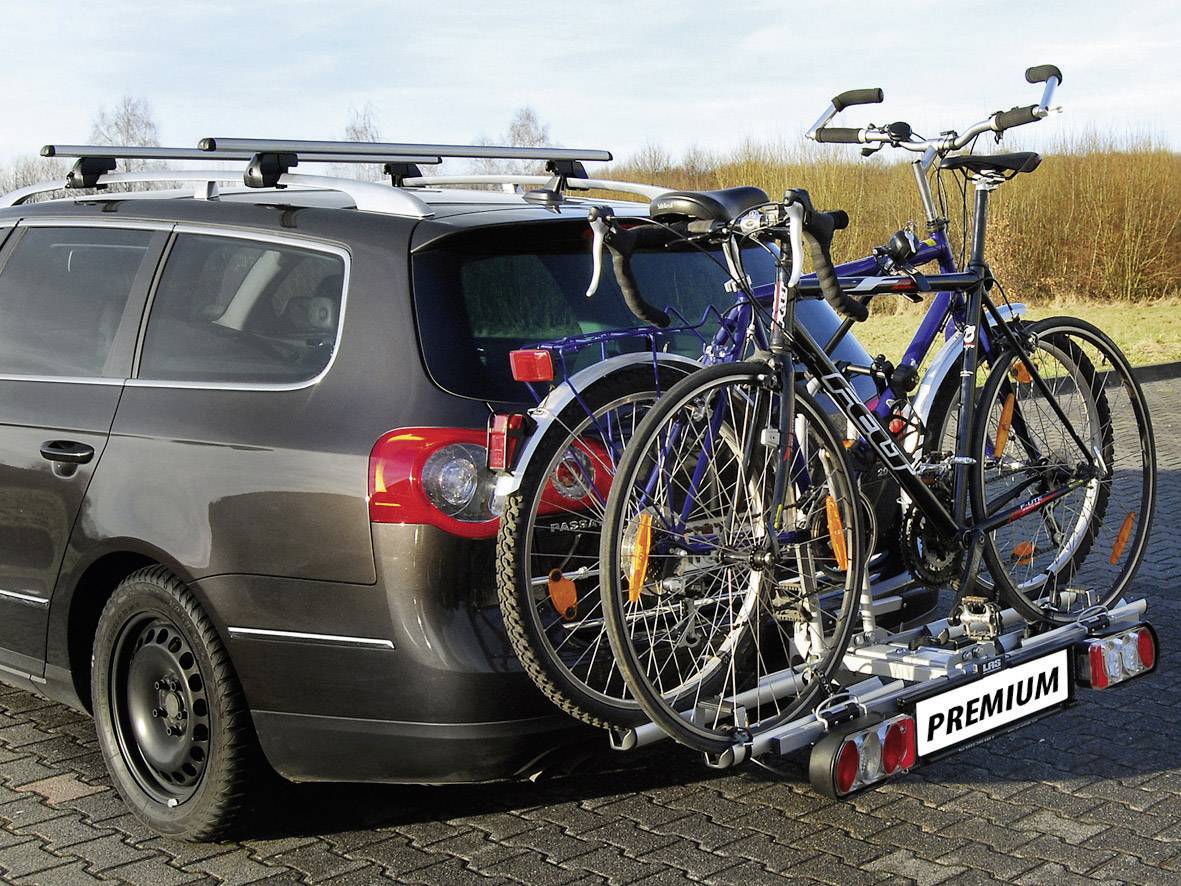Zilveren auto met twee fietsen op een fietsendrager aan de achterkant, geparkeerd op een geplaveid plein bij daglicht.