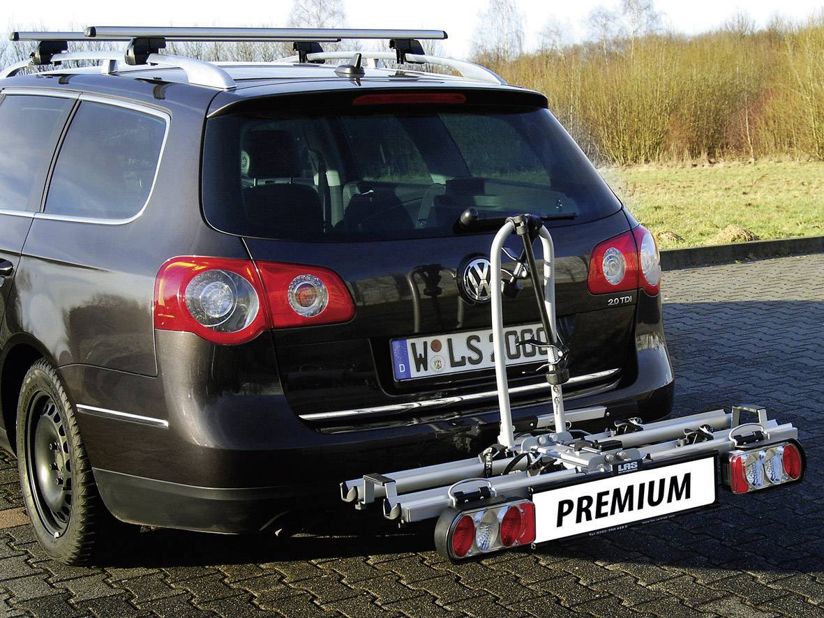 Een auto met een op de achterkant gemonteerde fietsendrager staat geparkeerd op een parkeerplaats, omgeven door gras en bomen op de achtergrond.
