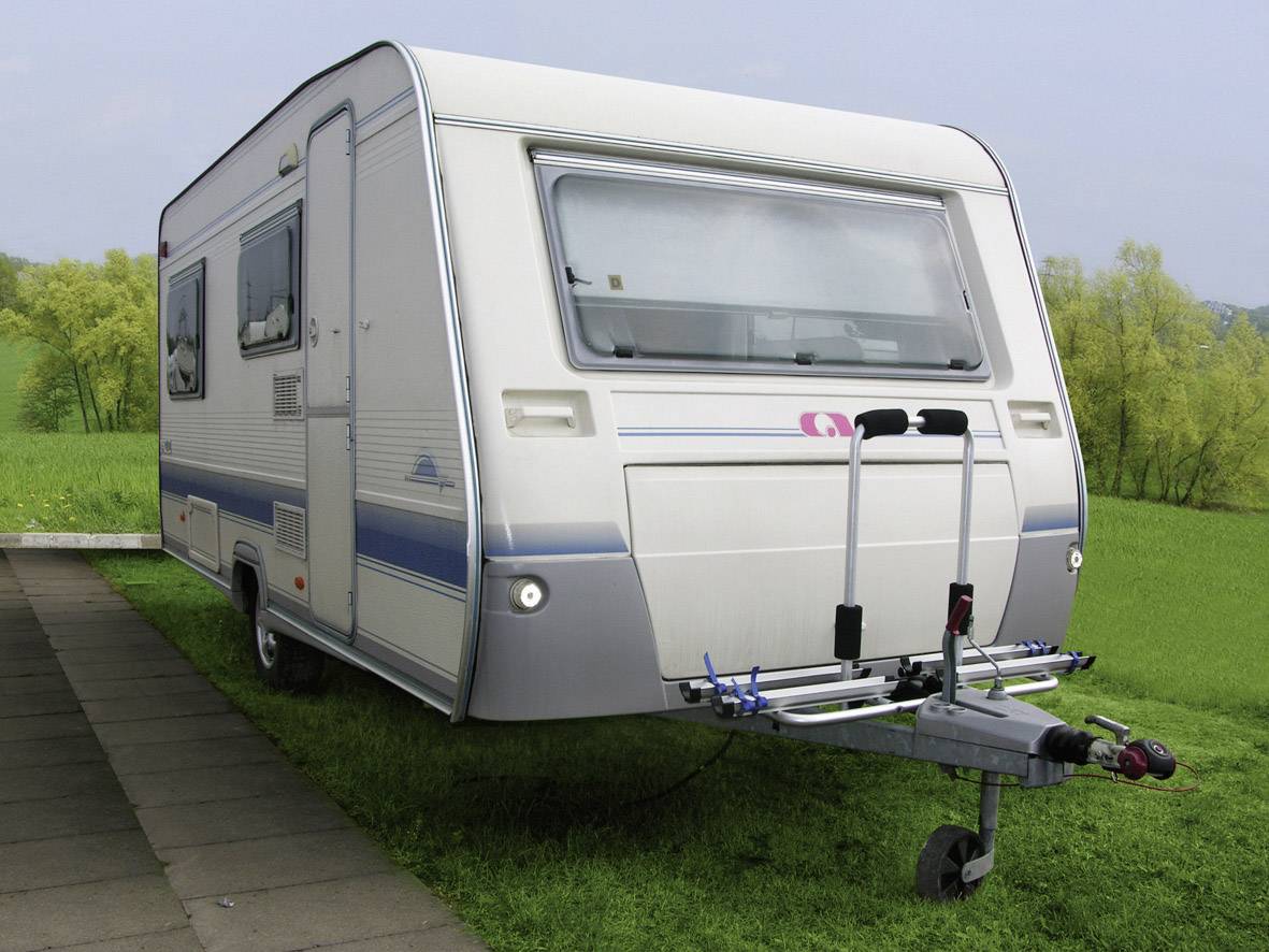 Witte caravan met fietsendrager, staat op een grasveld naast een bestrate weg, bomen op de achtergrond.