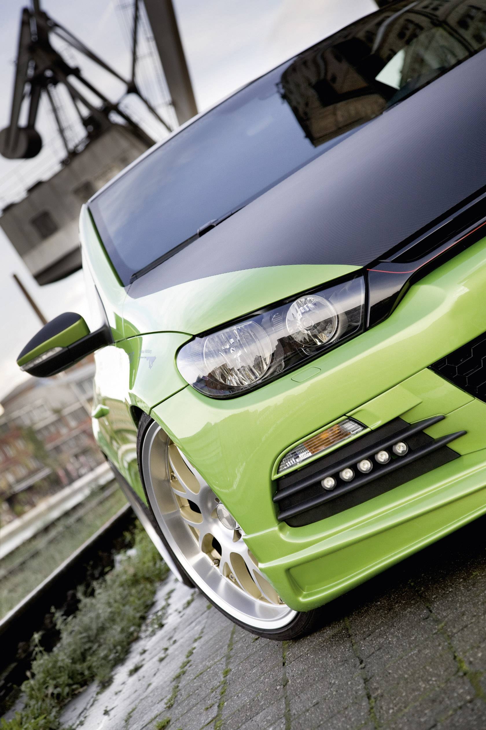 Groene auto met glanzende lak en een sportief design staat op een geplaveid wegdek naast spoorlijnen, met industriële structuren op de achtergrond.