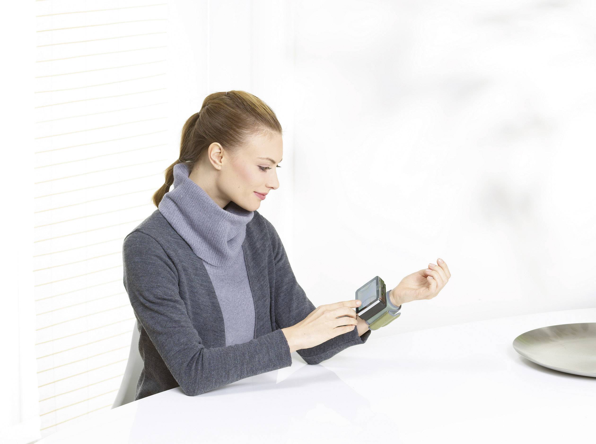 Een vrouw meet haar bloeddruk met een digitaal meetapparaat aan haar pols terwijl ze aan een tafel zit in een lichte ruimte.