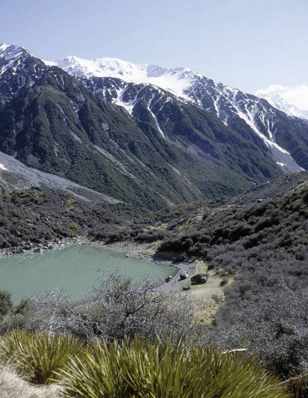 Een smaragdgroene meer ligt ingebed in een berglandschap met besneeuwde toppen onder een heldere, blauwe hemel.