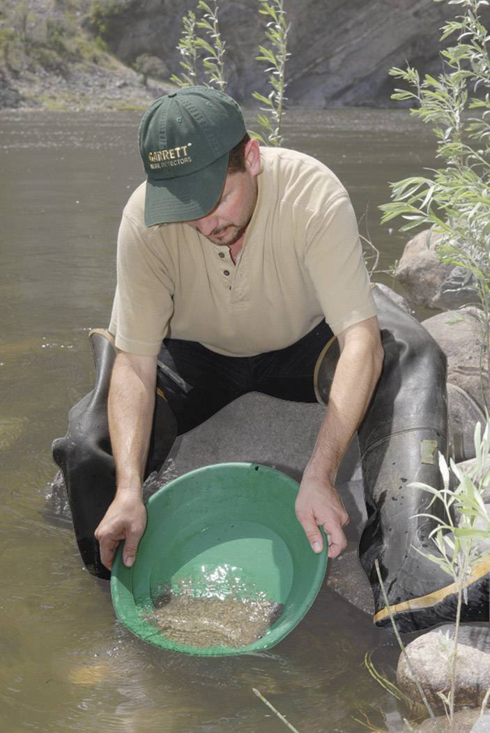 Een man in rubberlaarzen zoekt met een groene pan in een rivier naar goud. Hij leunt tegen een rots en kijkt geconcentreerd.