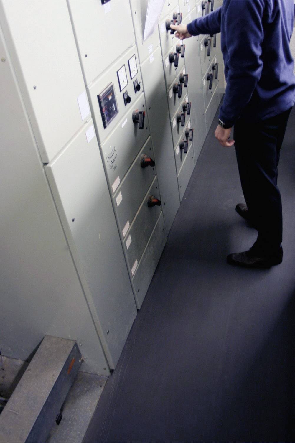 Een persoon bedient schakelaars aan een schakelkast in een technische ruimte. De wanden zijn uitgerust met bedieningselementen.