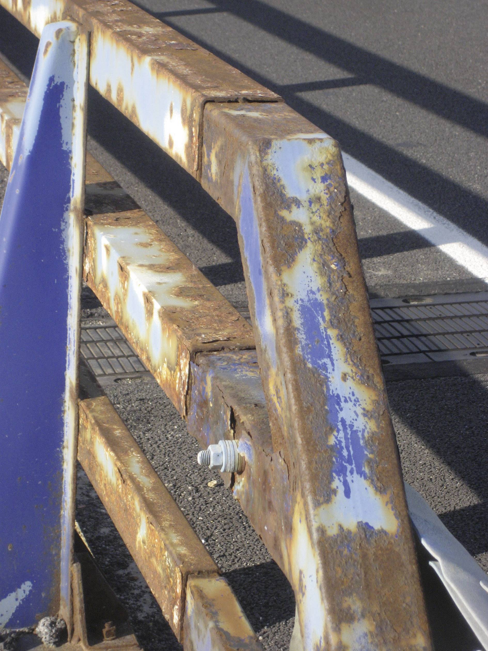 Verroest metalen leuningwerk met afbladderende blauwe verf op een straathoek, vertoont tekenen van verwering en verwaarlozing.