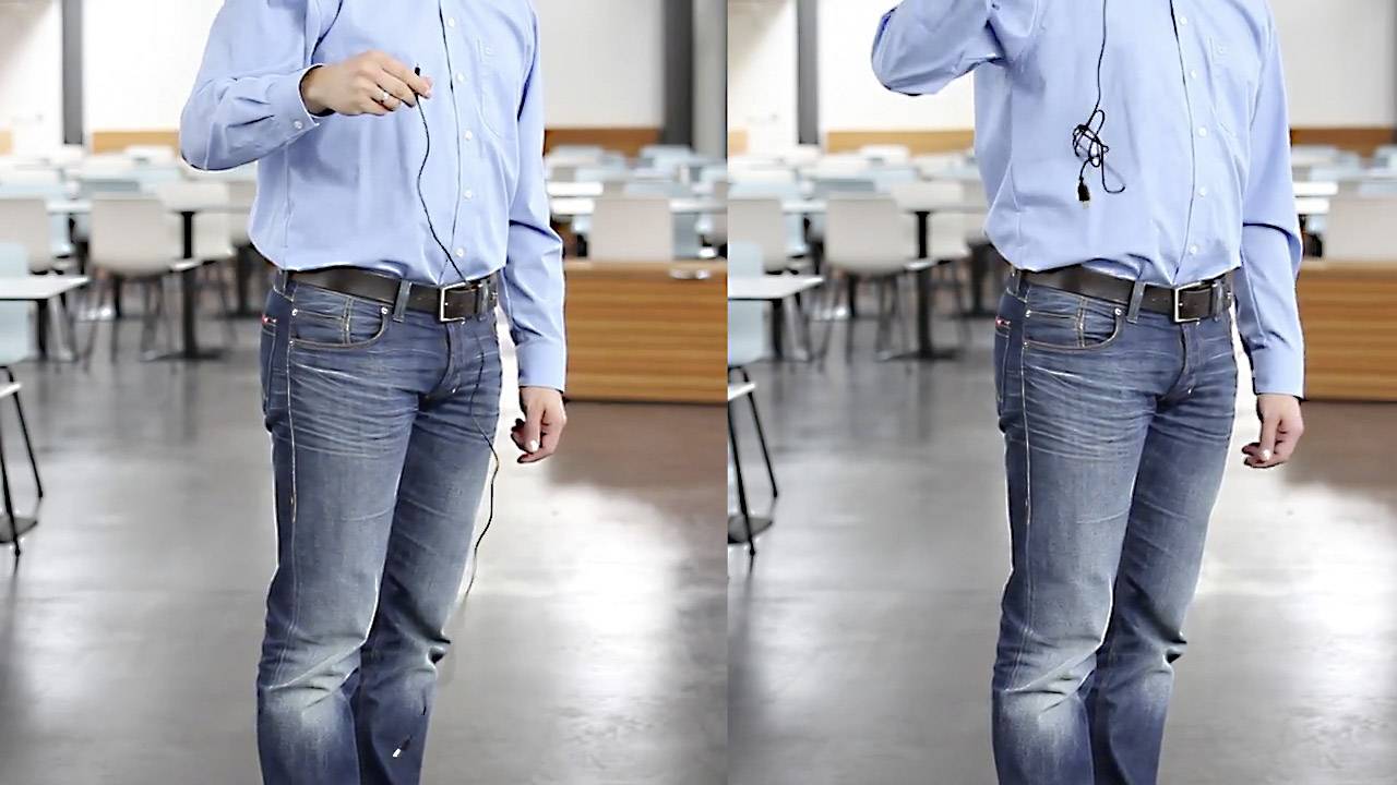 Een persoon in spijkerbroek en een blauw shirt staat in een lege klaslokaal. In de hand houdt hij/zij een koptelefoon.