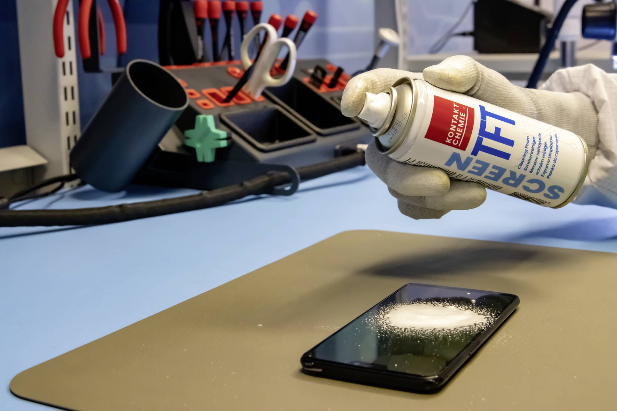 Osoba rozpyla środek czyszczący na smartfonie leżącym na stole. W tle widać narzędzia.