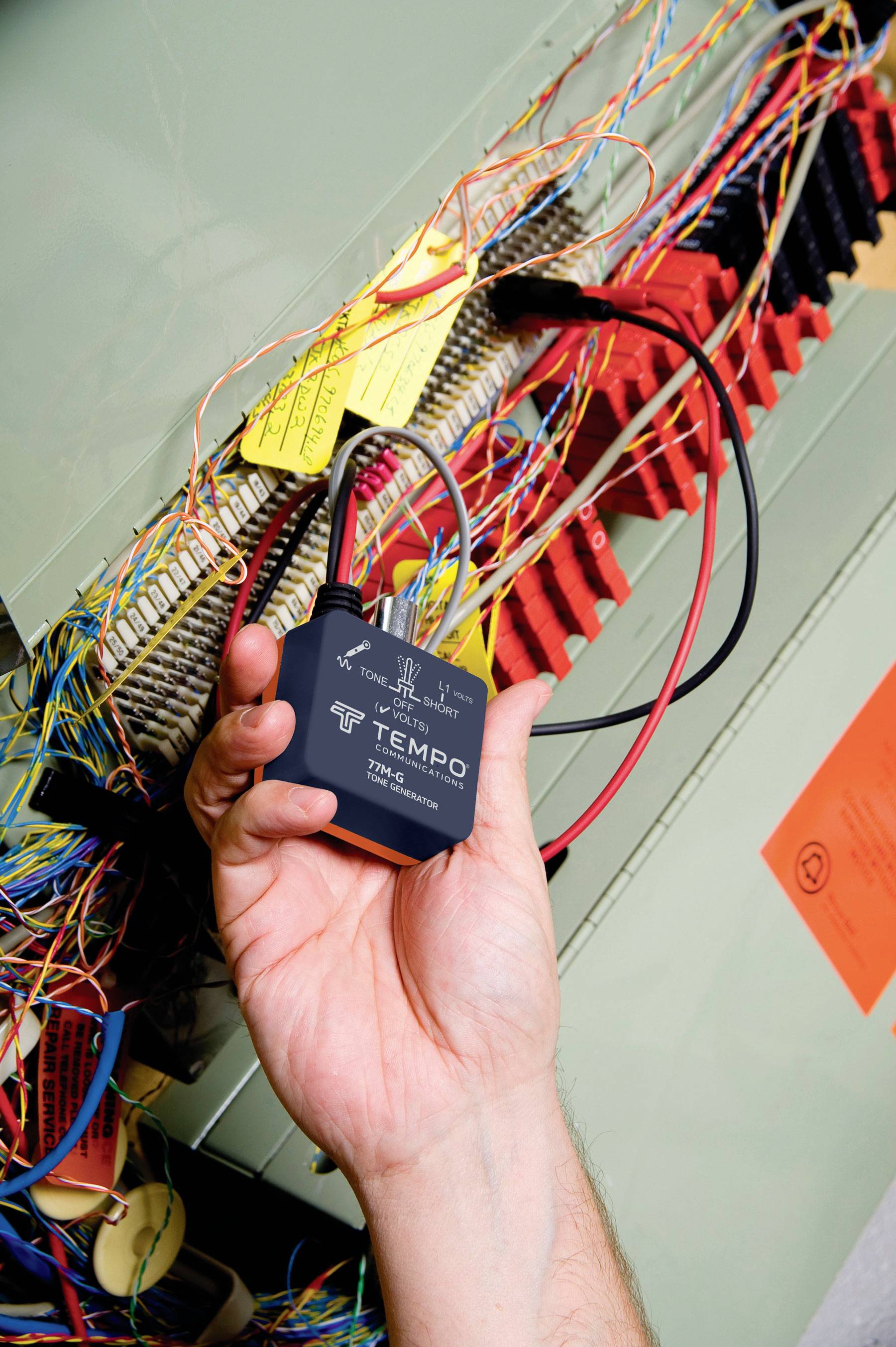 Dłoń trzyma elektryczne urządzenie pomiarowe z kablami wewnątrz szafy sterowniczej. Widoczne są kolorowe przewody i etykiety.