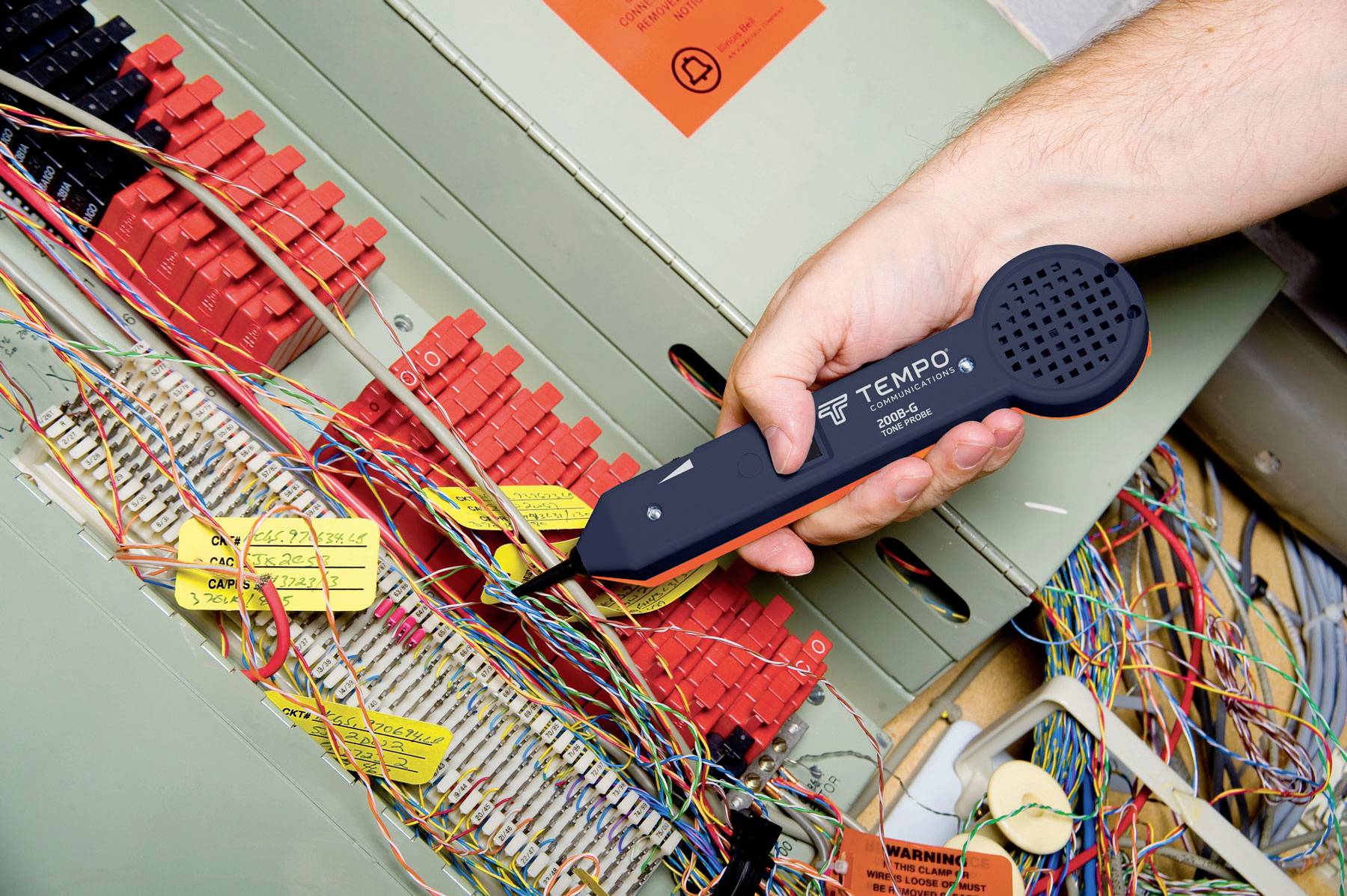 Dłoń trzyma urządzenie pomiarowe przy kablu telefonicznym wewnątrz szafki telekomunikacyjnej. Widoczne są różnokolorowe przewody.
